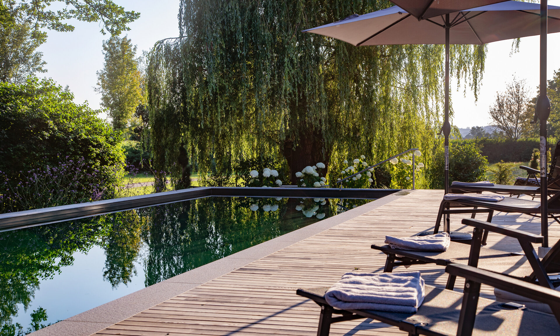 Holzterrasse mit stilvollen Stühlen und Schirmen, beim Naturpool des Hotel Quellenhof.