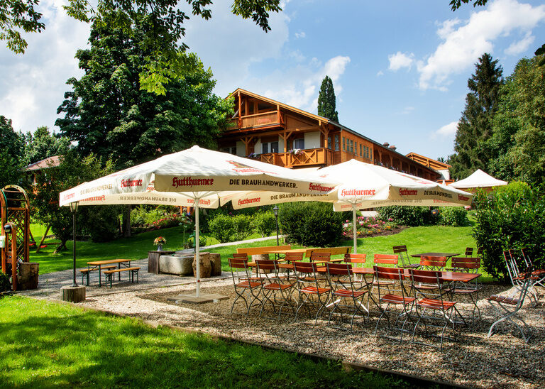 Ein Biergarten mit Stühlen und Tischen umgeben von grüner Wiese und Hotel Quellenhof im Hintergrund.