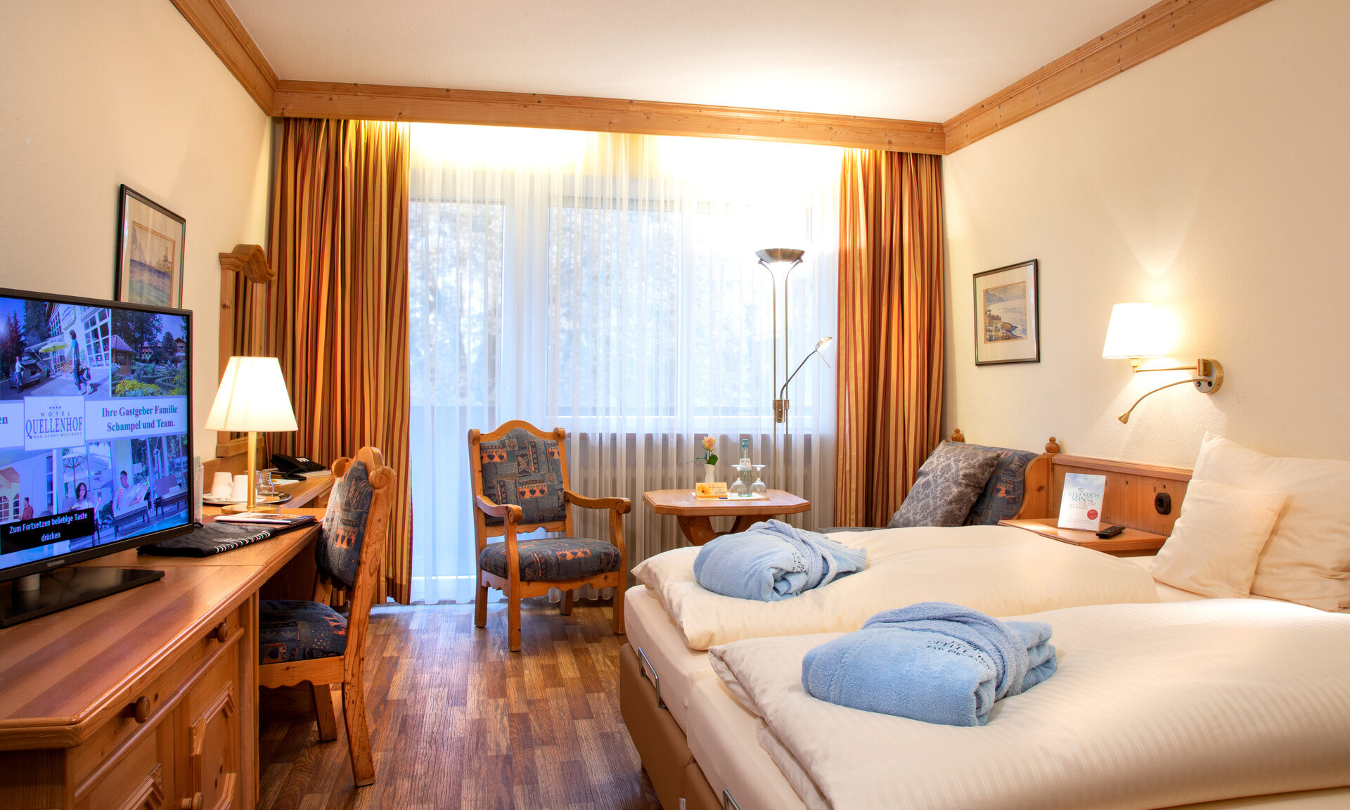 Eine wohnliche Atmosphäre im Schlafzimmer des Hotel Quellenhof mit großem Fenster, Sitzgelegenheit und einem Fernseher.