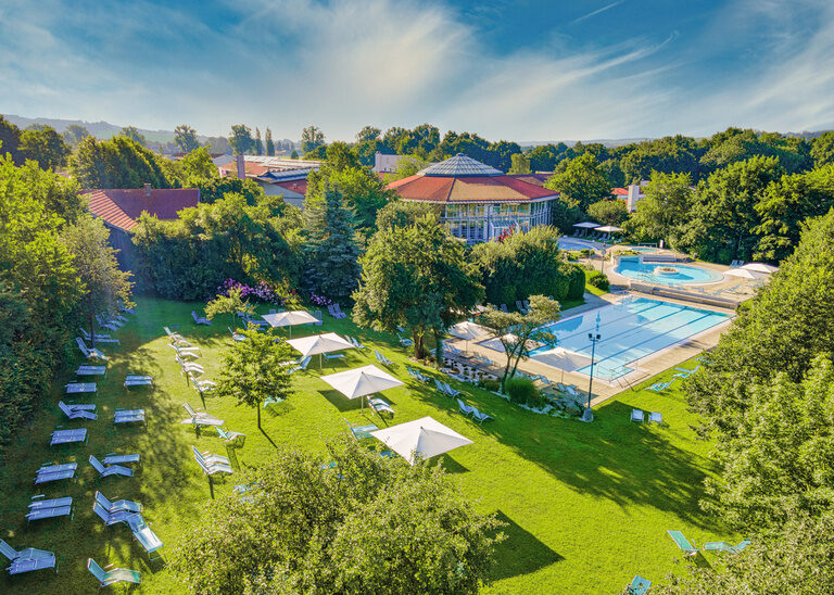 Blick auf die grüne Liegewiese der Rottal Terme mit Liegen und Schirmen sowie Becken im Hintergrund an einem schönen sommerlichen Tag.