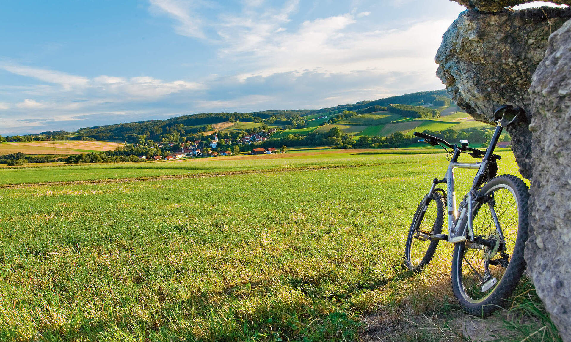 Ein Mountainbike lehnt an einer steinernen Mauer mit malerischer Wiese im Rottal im Hintergrund.