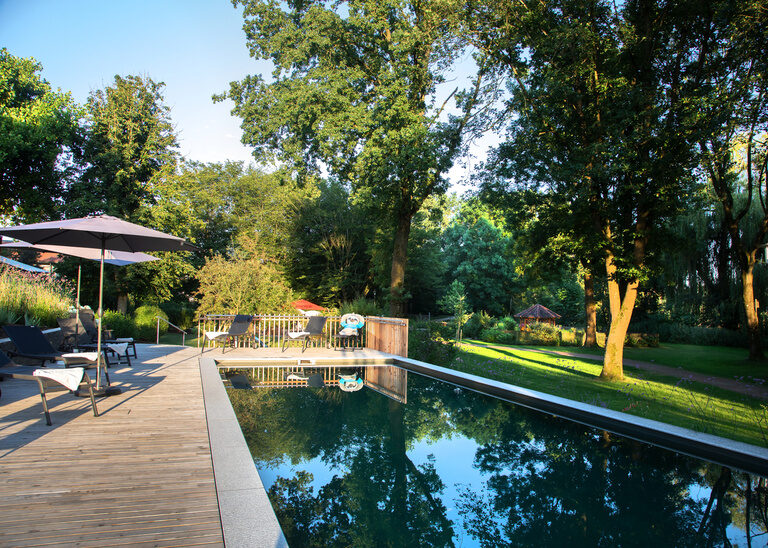 Der Naturpool des Hotel Quellenhof mit Sonnenterrasse, auf der Sonnenliegen und Sonnenschirme stehen.